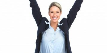 Woman sitting on the ground in front of her laptop with her arms up in celebration while she smiles at the camera