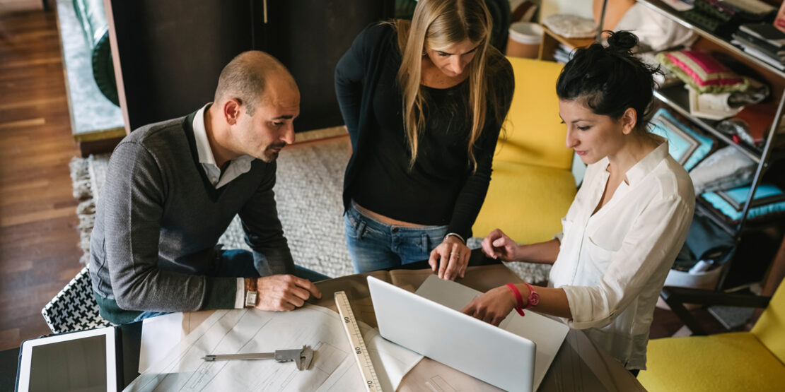 3 designers looking at a laptop at a drawing table with blueprints in front of them