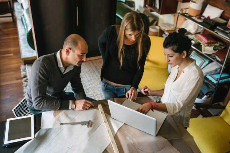 3 designers looking at a laptop at a drawing table with blueprints in front of them