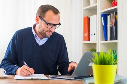 man writing on a clipboard as he scrolls on his laptop