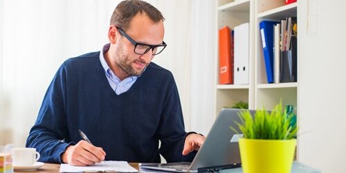 man writing on a clipboard as he scrolls on his laptop