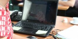 woman writing on a piece of paper at her laptop as people behind her talk and work