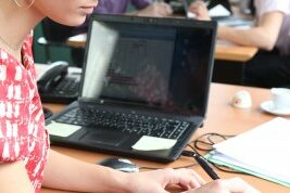 woman writing on a piece of paper at her laptop as people behind her talk and work