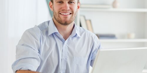 man smiling at the camera as he works on a laptop