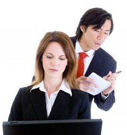 woman working on her computer with a man looking over her shoulder writing something down on a notepad
