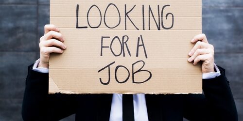 Man holding up a cardboard sign saying he is looking for a job in front of his face as he is dressed in a suit