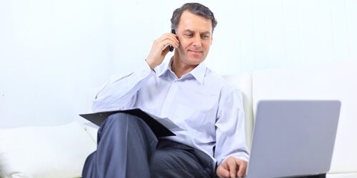 man on his phone working on his laptop on a couch