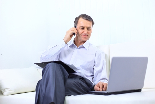 man on his phone working on his laptop on a couch