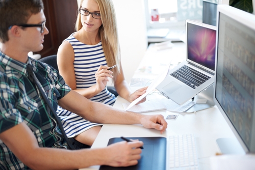 Coworkers sitting at a desk in front of their computers discussing