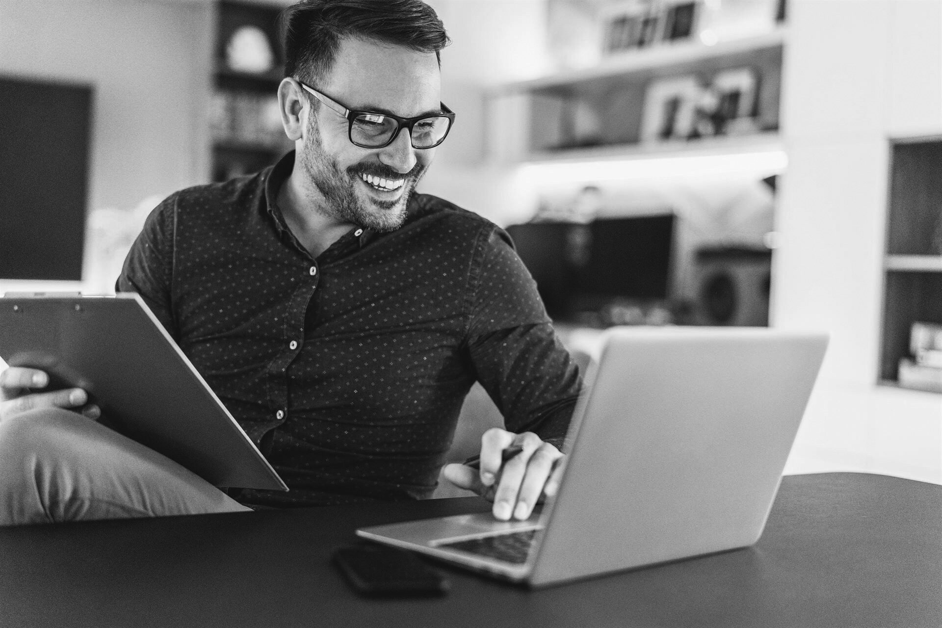 Man sitting at a desk staring at his computer smiling, he is holding a clipboard in one hand and a pen in his other.