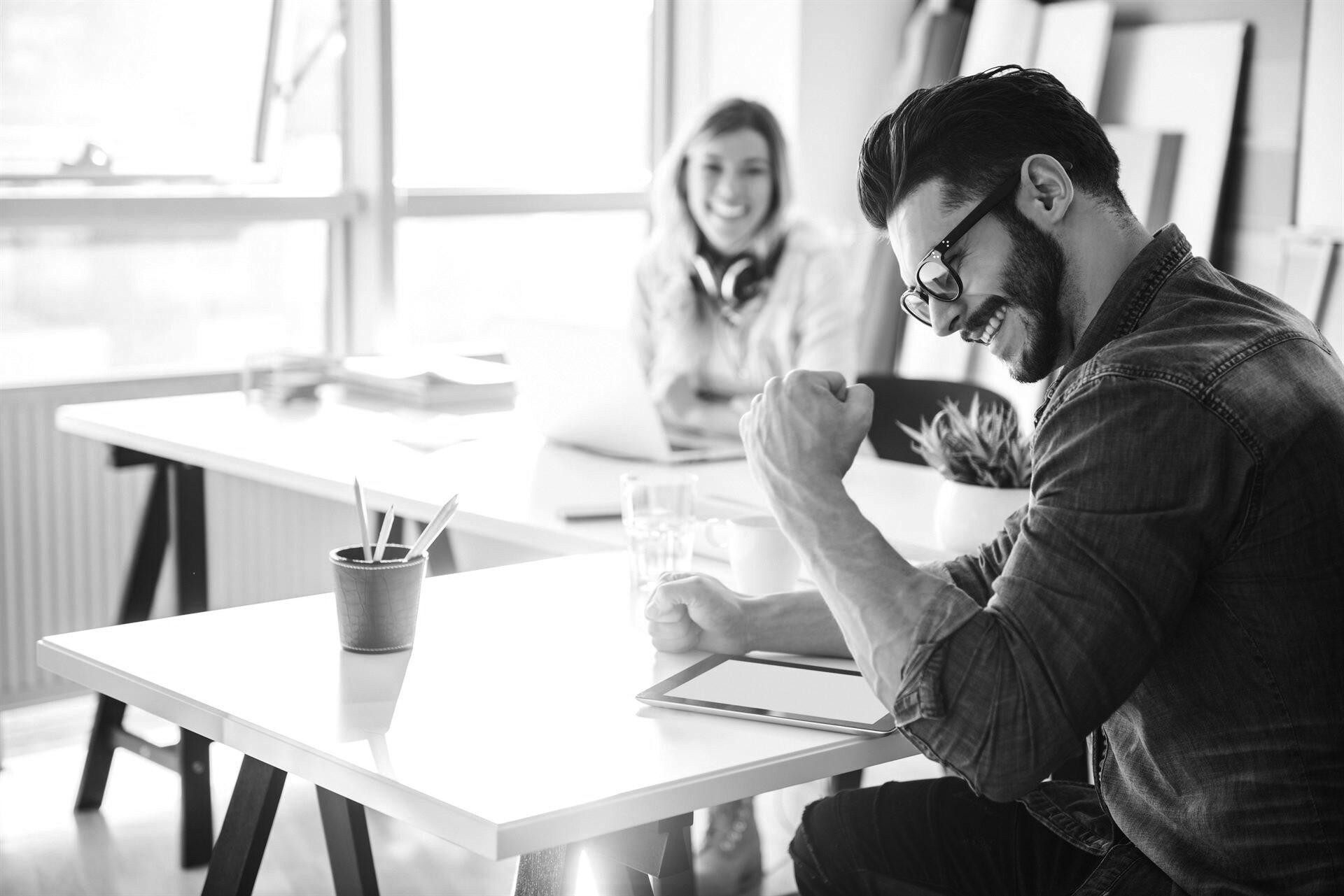 Man sitting at a communal desk staring at a tablet as he pumps his fist in excitement. A woman is sitting at the same desk in the background smiling at him as she works on a laptop. She is out of focus.