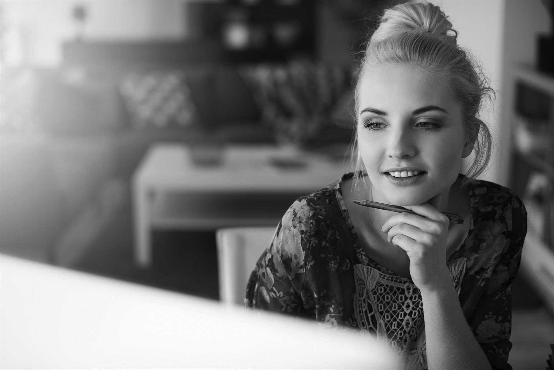 Close up of a woman sitting at a desk in her living room with her hand under her chin. The computer is blurred in front and she is holding a pen.