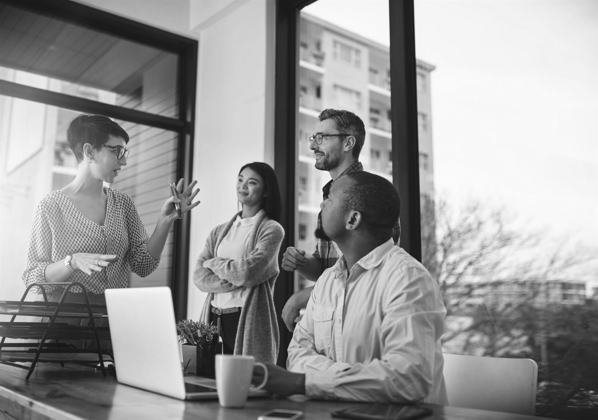 Group of coworkers standing by ones desk as they engage in a discussion in front of a large glass window