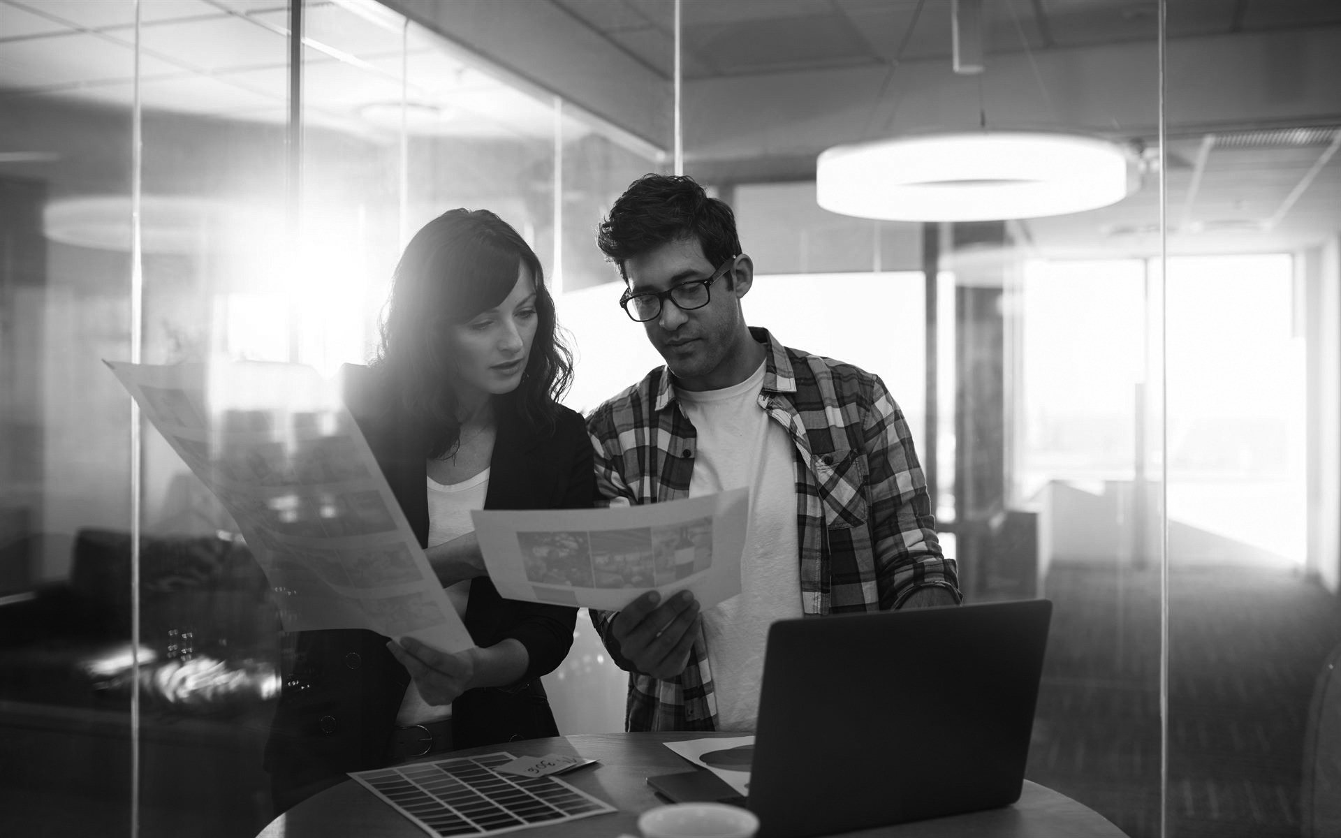 2 coworkers looking at images and color swatches at a small table in the middle of a glass office with a laptop sitting on the table as well.